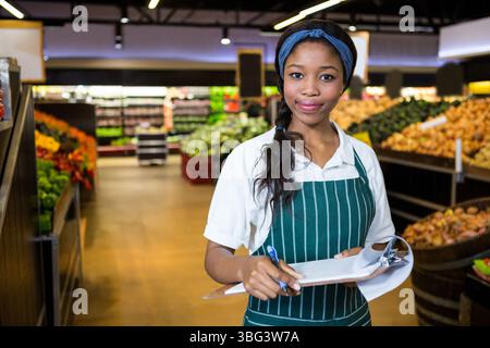 Femme afro-américaine portant un tablier rayé vérifiant les produits avec une planche à pince dans l'allée du supermarché Banque D'Images
