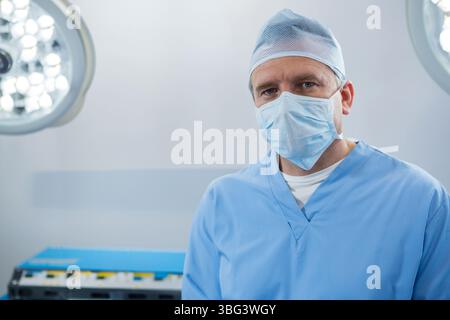 Chirurgien masculin senior debout en gommage en salle d'opération sous lumières chirurgicales près de la table d'instruments Banque D'Images