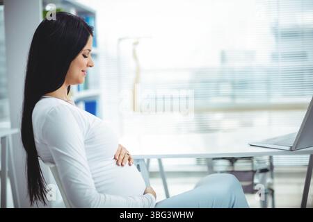 Femme enceinte assise au bureau près des stores de fenêtre berçant la bosse, regardant vers le bas l'ordinateur portable, espace de copie Banque D'Images