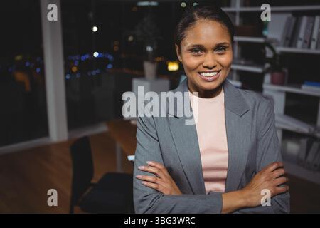 Femme d'affaires debout dans un bureau moderne la nuit avec bureau, étagères avec plante et skyline de la ville Banque D'Images