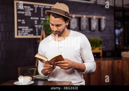Homme barbu lisant livre de poche par tasse à café sur table dans le café avec menu tableau noir Banque D'Images