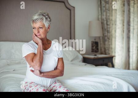 Femme âgée reposant la joue sur la main et croisant les bras sur le lit avec la literie blanche à côté de la lampe Banque D'Images