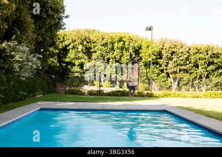 Femme dans les années 20 portant un maillot de bain deux pièces vert fluo faisant du saut de ligne au-dessus de la terrasse de la piscine en pierre de l'arrière-cour Banque D'Images