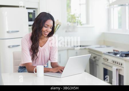 Femme afro-américaine souriante tout en tapant sur ordinateur portable au comptoir de cuisine avec tasse et fleurs Banque D'Images