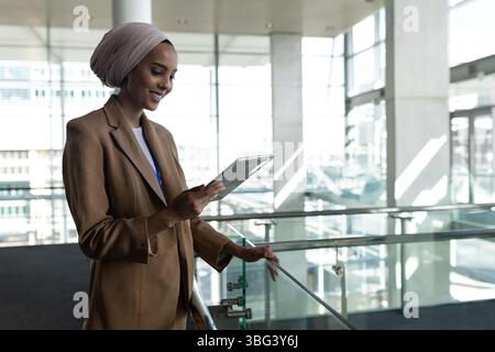 Femme d'affaires portant blazer tan et hijab s'appuyant sur la balustrade en verre et utilisant la tablette dans l'atrium Banque D'Images