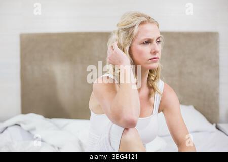 Femme adulte moyenne portant des vêtements de nuit blancs assis sur le lit avec des draps dans la chambre, regardant à droite Banque D'Images