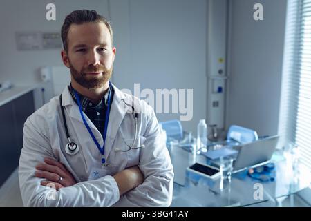 Médecin masculin debout avec les bras croisés dans le bureau de la clinique à côté de l'ordinateur portable portant manteau et stéthoscope Banque D'Images