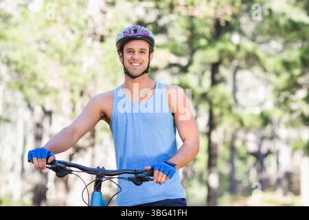 Cycliste masculin debout souriant sur un sentier forestier ensoleillé tenant le guidon de vélo portant un casque Banque D'Images