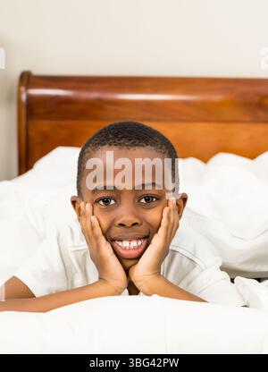 Garçon afro-américain allongé sur le lit avec des draps blancs et des oreillers dans la chambre, se reposant et souriant Banque D'Images