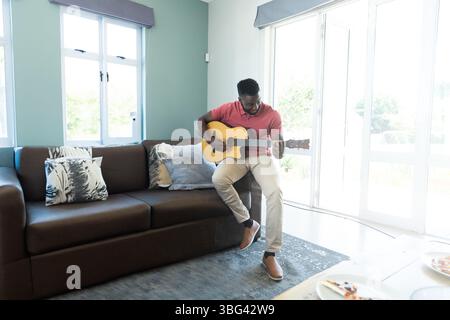 Homme afro-américain strumming guitare acoustique sur canapé en cuir dans le salon à côté des tranches de pizza Banque D'Images
