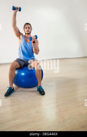 Homme en tenue de fitness assis sur un ballon d'exercice bleu soulevant un haltère au-dessus de la tête dans un studio de fitness Banque D'Images