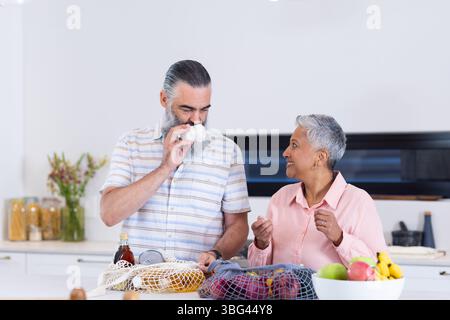 Couple senior diversifié déballage de l'épicerie derrière l'îlot de cuisine avec des sacs de produits en maille Banque D'Images