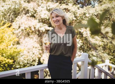 Femme debout sur le pont en bois dans la cour arrière tenant le verre de vin et s'appuyant sur la balustrade Banque D'Images