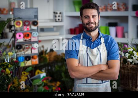 Fleuriste masculin debout bras croisés dans un magasin de fleurs montrant des bobines de ruban et des fleurs coupées, espace de copie Banque D'Images