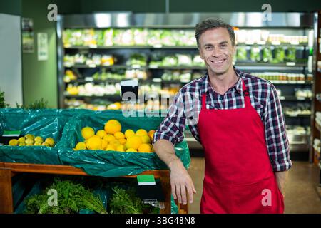 Homme d'âge moyen dans un tablier appuyé sur le bac de l'allée des produits du supermarché montrant des citrons avec une affiche de prix Banque D'Images