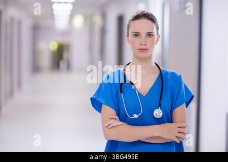 Infirmière debout avec les bras croisés dans le couloir de l'hôpital portant des gommages bleus et stéthoscope Banque D'Images