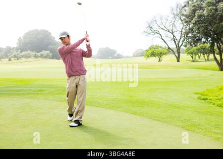 Homme asiatique debout sur le vert de mise tenant le fer de golf pendant la journée ensoleillée sur le fairway bordé d'arbres Banque D'Images