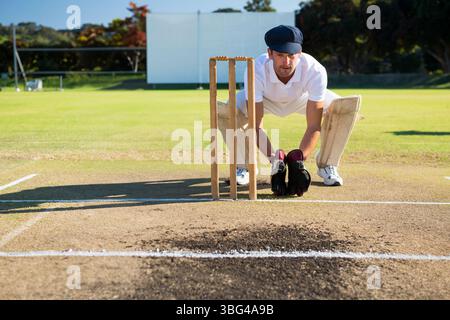 Gardien de guichet masculin portant un kit blanc accroupi derrière des souches sur des coussins de gants de terrain avec écran visuel Banque D'Images