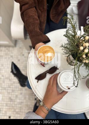 Mains tenant un latte avec des œuvres d'art en mousse en forme de coeur au-dessus d'une paire de lunettes de soleil et de verdure sur une table de café en marbre. Banque D'Images