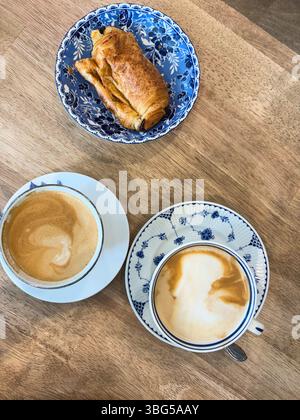 Un croissant au chocolat dans un bol floral bleu se trouve à côté de deux tasses de café sur une table en bois. Banque D'Images