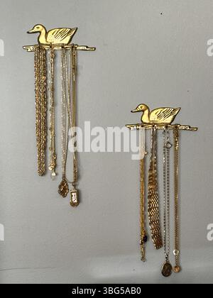 Les cintres muraux de canard décoratifs en laiton affichent soigneusement une variété de colliers et de chaînes en or sur un mur peint gris. Banque D'Images