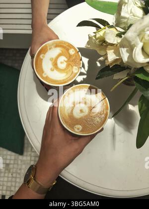 Deux tasses en papier de café avec latte art en forme d'ours sont assis sur une table blanche à côté d'un bouquet de roses blanches. Banque D'Images
