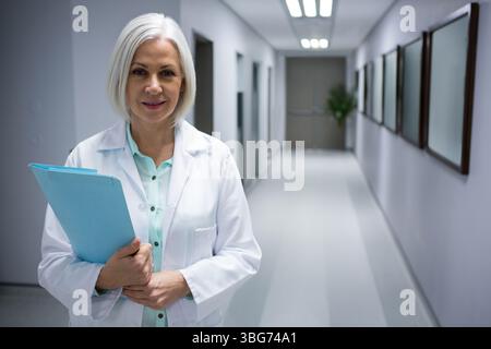 Médecin femme d'âge moyen portant une blouse de laboratoire et tenant un dossier bleu dans le couloir de l'hôpital, espace de copie Banque D'Images