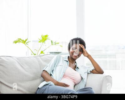 Femme enceinte afro-américaine assise sur un canapé gris par la fenêtre touchant le ventre, espace de copie Banque D'Images