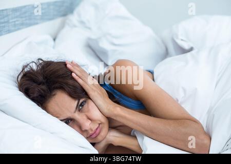 Femme pressant les mains sur les oreilles dans le débardeur bleu sous couette blanche sur le lit dans la chambre Banque D'Images