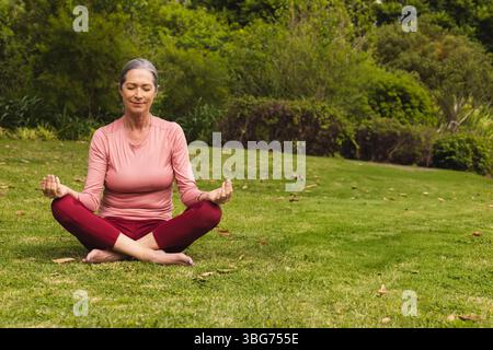 Femme senior méditant sur la pelouse herbeuse dans le jardin portant un haut athlétique rose et un pantalon Bordeaux Banque D'Images