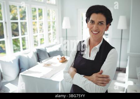 Femme debout souriant dans le coin lumineux du petit déjeuner portant tablier noir sur chemise blanche, espace de copie Banque D'Images