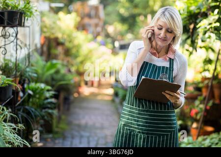 Femme senior portant tablier à l'aide d'une presse-papiers et d'un téléphone, vérifiant les plantes dans l'allée du jardin, espace de copie Banque D'Images