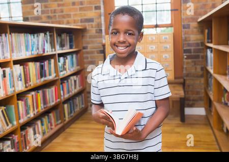 Garçon afro-américain tenant un livre ouvert, souriant et debout près du banc de bibliothèque et du catalogue de cartes Banque D'Images