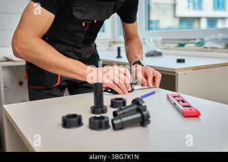 Homme mesurant la partie de meuble lors de l'assemblage des armoires de cuisine, à l'aide de ruban à mesurer, d'outils et de pieds de meuble en plastique Banque D'Images