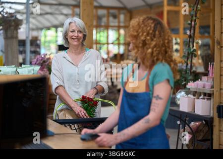 Femme senior achetant des fleurs à la caisse du centre de jardin : payer en utilisant la technologie sans contact Banque D'Images
