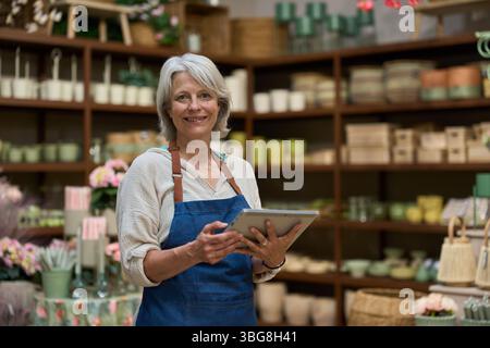 Vendeuse senior utilisant la tablette numérique dans le magasin de fleurs : gestion des stocks avec la technologie Banque D'Images