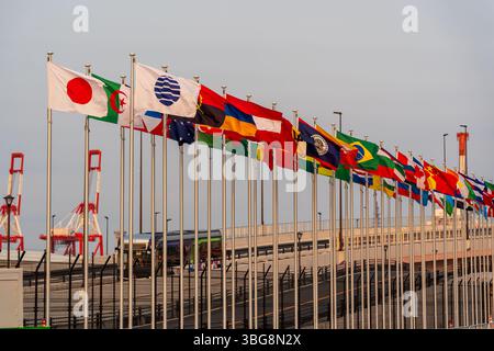 Les drapeaux internationaux flottent majestueusement contre un ciel crépusculaire clair à l'entrée de l'Expo 2025 Osaka Kansai pendant le coucher du soleil. Osaka, Japon Banque D'Images