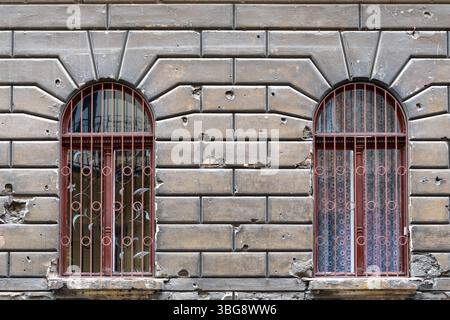 La façade d'un ancien bâtiment avec deux fenêtres cintrées décorées de treillis en fer forgé. Murs en pierre avec traces visibles de dommages causés par des balles Banque D'Images