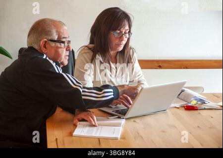 Un homme âgé et une femme adulte sont assis à une table en bois, travaillant ensemble tout en étant concentrés sur un écran d'ordinateur portable. Autour d'eux se trouvent des documents, des stylos, un Banque D'Images