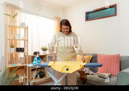 Femme faisant des vêtements travaux ménagers repassage corvée Maison ménage asiatique appareil féminin, Lady souriant tout en repassant et pliant blanchisserie proprement dans un l lumineux Banque D'Images