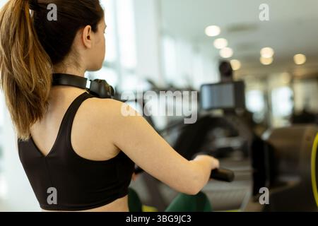 Une jeune femme dévouée s'entraîne sur un rameur, se concentrant intensément dans un environnement de gymnastique dynamique et moderne. Banque D'Images