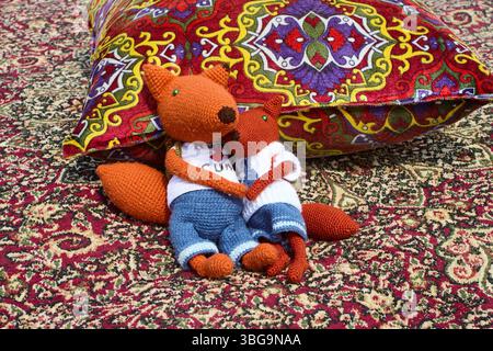 Deux jouets de renard en tricot doux, habillés de vêtements et avec des sacs à dos, blottis l'un à l'autre sur un tapis oriental coloré à côté d'un oreiller de jet, créant Banque D'Images