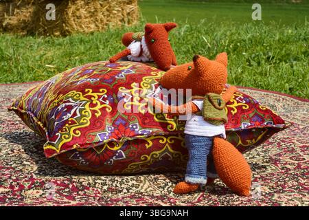 Deux jouets de renard en tricot doux, habillés de vêtements et avec des sacs à dos, blottis l'un à l'autre sur un tapis oriental coloré à côté d'un oreiller de jet, créant Banque D'Images