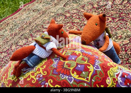 Deux jouets de renard en tricot doux, habillés de vêtements et avec des sacs à dos, blottis l'un à l'autre sur un tapis oriental coloré à côté d'un oreiller de jet, créant Banque D'Images