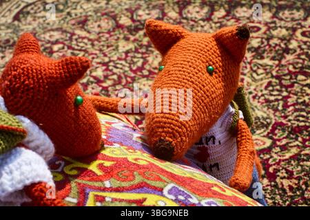 Deux jouets de renard en tricot doux, habillés de vêtements et avec des sacs à dos, blottis l'un à l'autre sur un tapis oriental coloré à côté d'un oreiller de jet, créant Banque D'Images