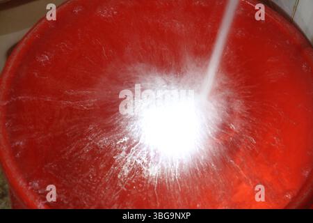 Seau rouge rempli d'eau provenant d'un courant à haute pression. Le courant d'eau frappe le centre du récipient, provoquant des éclaboussures et des turbulences. Banque D'Images