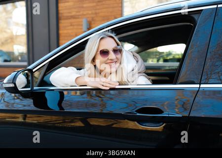 Femme blonde heureuse portant des lunettes de soleil élégantes, conduisant une voiture moderne avec un grand sourire, profitant de la liberté de la route ouverte Banque D'Images