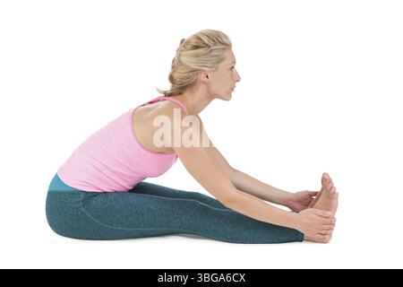 Vue latérale de profil complet du corps d'une femme blonde d'âge moyen dans la pose de yoga assise se pencher vers l'avant (Paschimothanasana) sur un fond blanc Banque D'Images