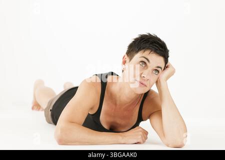 Vue complète du corps d'une femme d'âge moyen aux cheveux noirs pieds nus en short et haut noir couché sur son ventre sur un fond blanc regardant rêvement u Banque D'Images