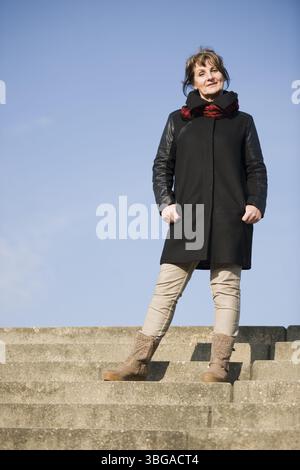 Portrait frontal pleine longueur d'une femme mature dans une veste en cuir noir debout sur un escalier par en-dessous Banque D'Images
